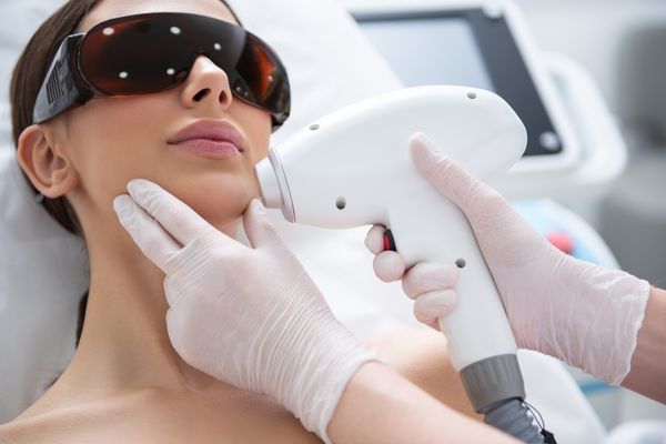 A beautiful woman with black safety goggles on her eyes receiving laser hair removal treatment on her chin from a professional technician