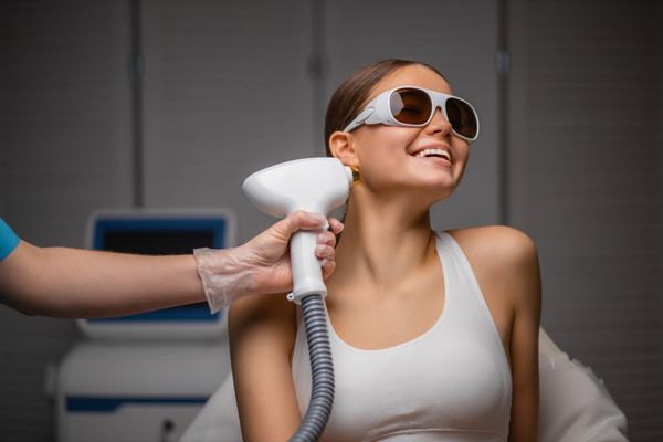 A smiling young woman receiving laser hair removal treatment on her sideburns from a professional technician while wearing safety goggles
